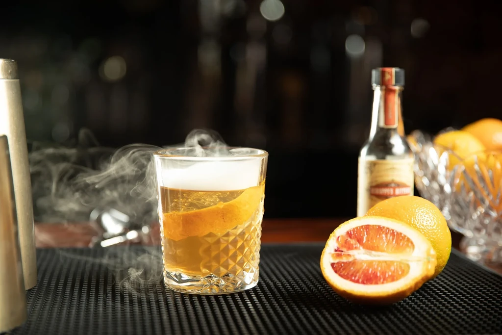 Faceted glass with smoking amber cocktail, orange peel, and blood oranges.