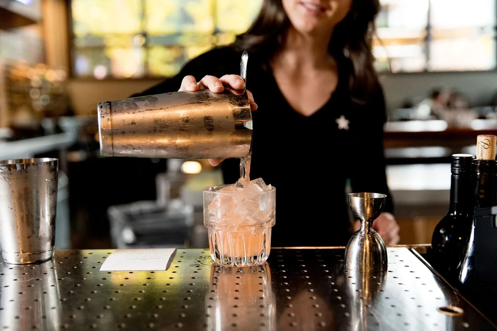 Bartender pouring a pink cocktail from a shaker into an iced glass.