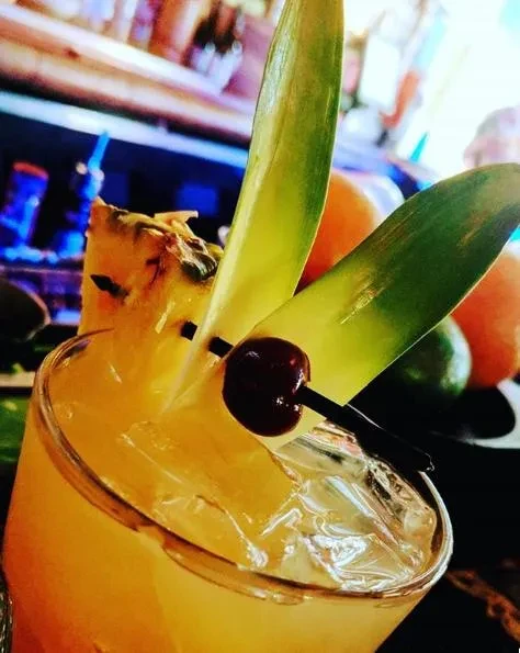 Yellow-orange cocktail with pineapple, cherry, ice in a bar setting.
