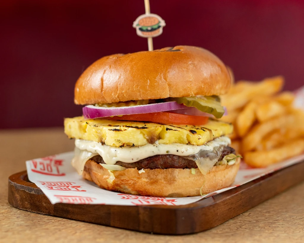 Gourmet burger with pineapple, cheese, and fries on a wooden board.