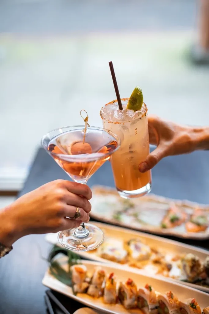 Two hands toast a martini and a tall iced cocktail.