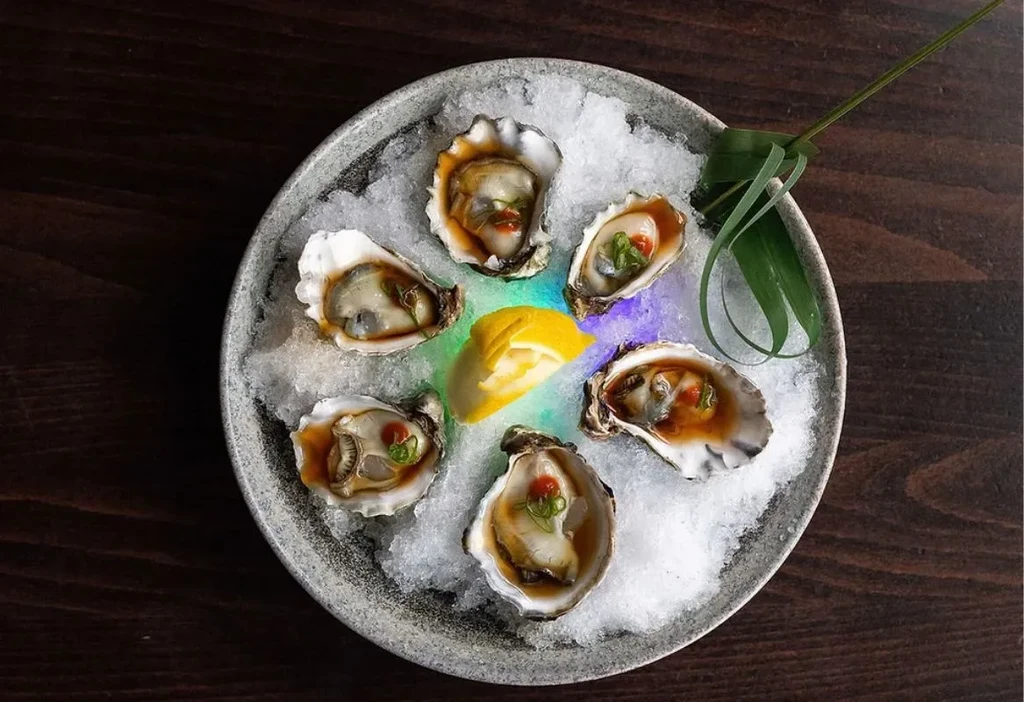 Six garnished oysters on crushed ice with lemon on a grey plate.