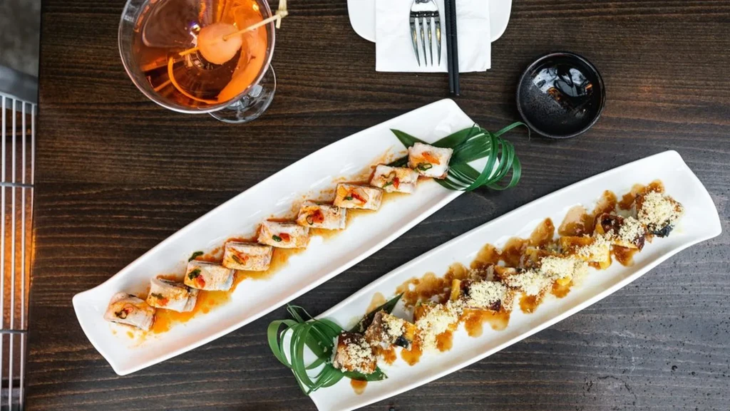 Overhead view: Two sushi plates, cocktail, chopsticks on a dark wooden table.