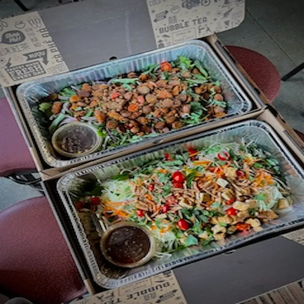 Two open takeout boxes, foil trays with varied salads and sauce.