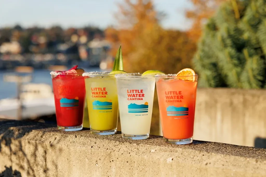 Four colorful Little Water Cantina drinks garnished by a sunny waterfront.