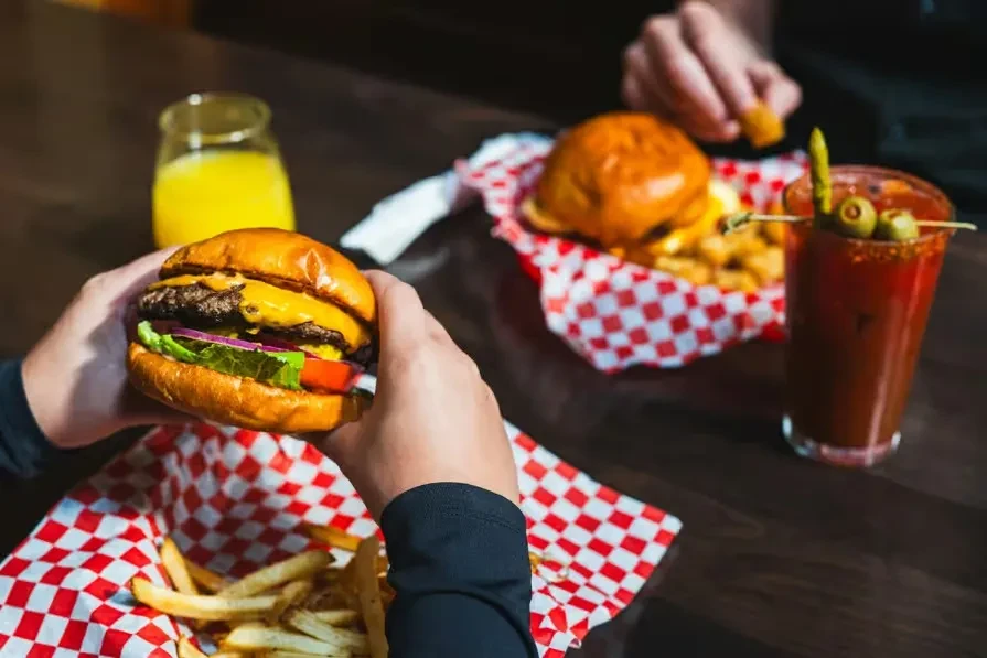 Hands hold a cheeseburger, fries, juice, and cocktail on a table.