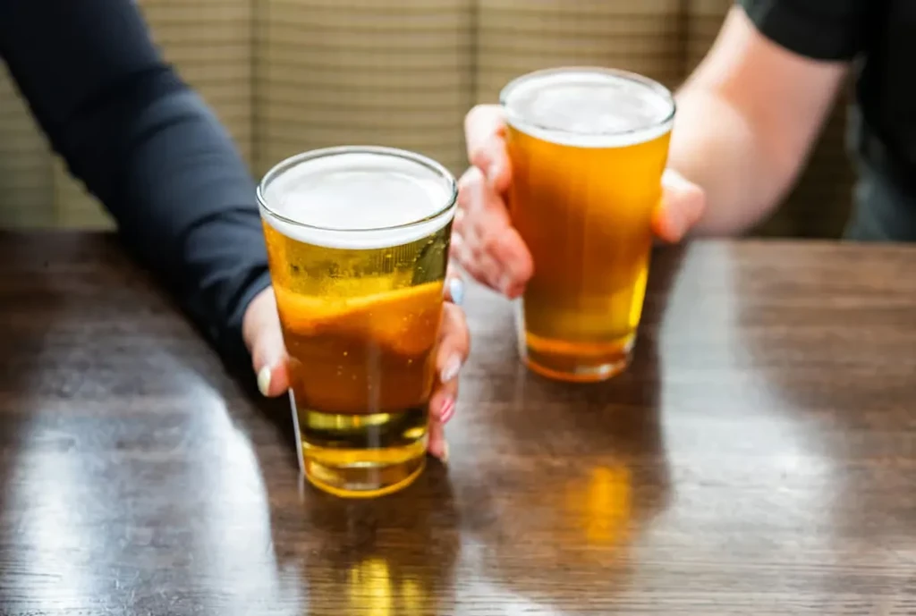 Two hands hold two foamy beers on a polished wooden table.