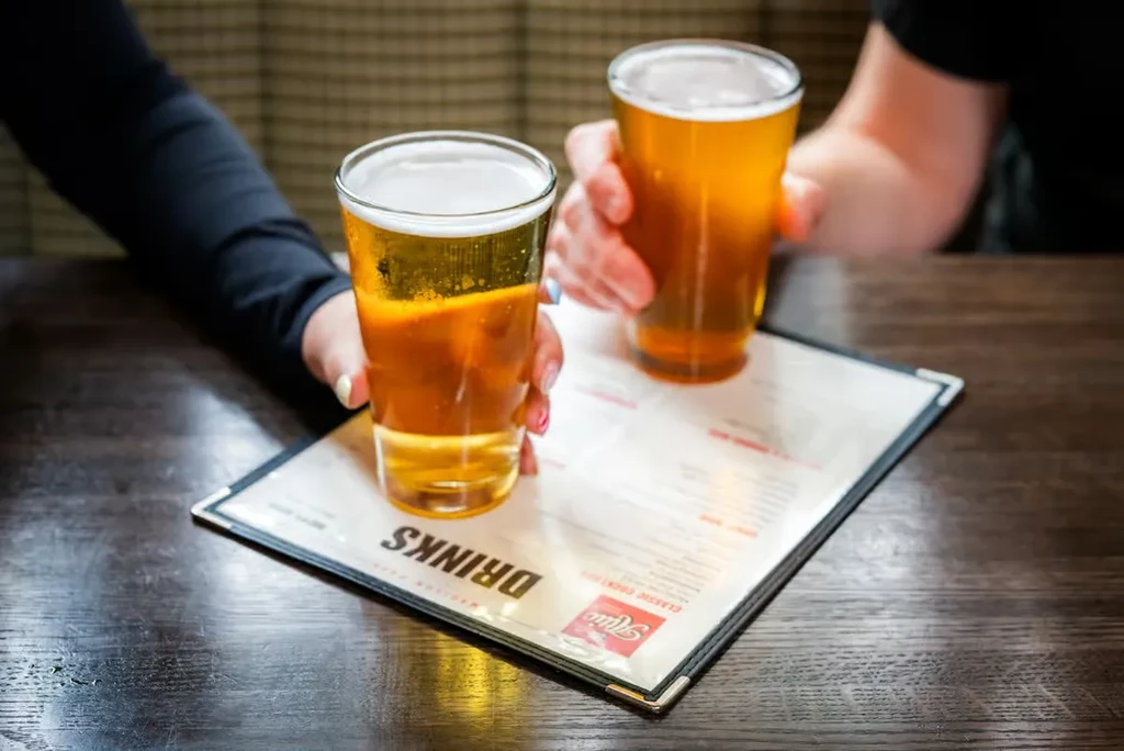 Two hands hold pint glasses of amber beer above a 'DRINKS' menu.