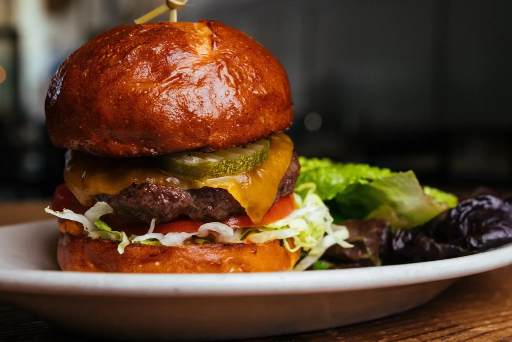 Stacked gourmet burger with cheese, pickles, tomato, lettuce, and side salad.