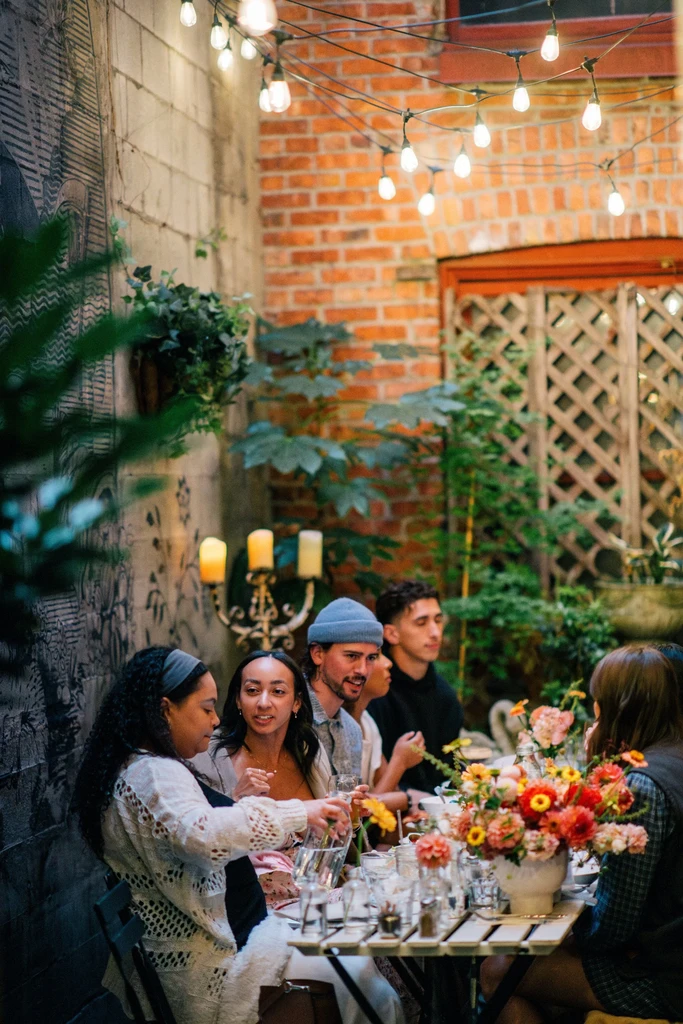 Group of people dining outdoors with flowers, drinks, and string lights.
