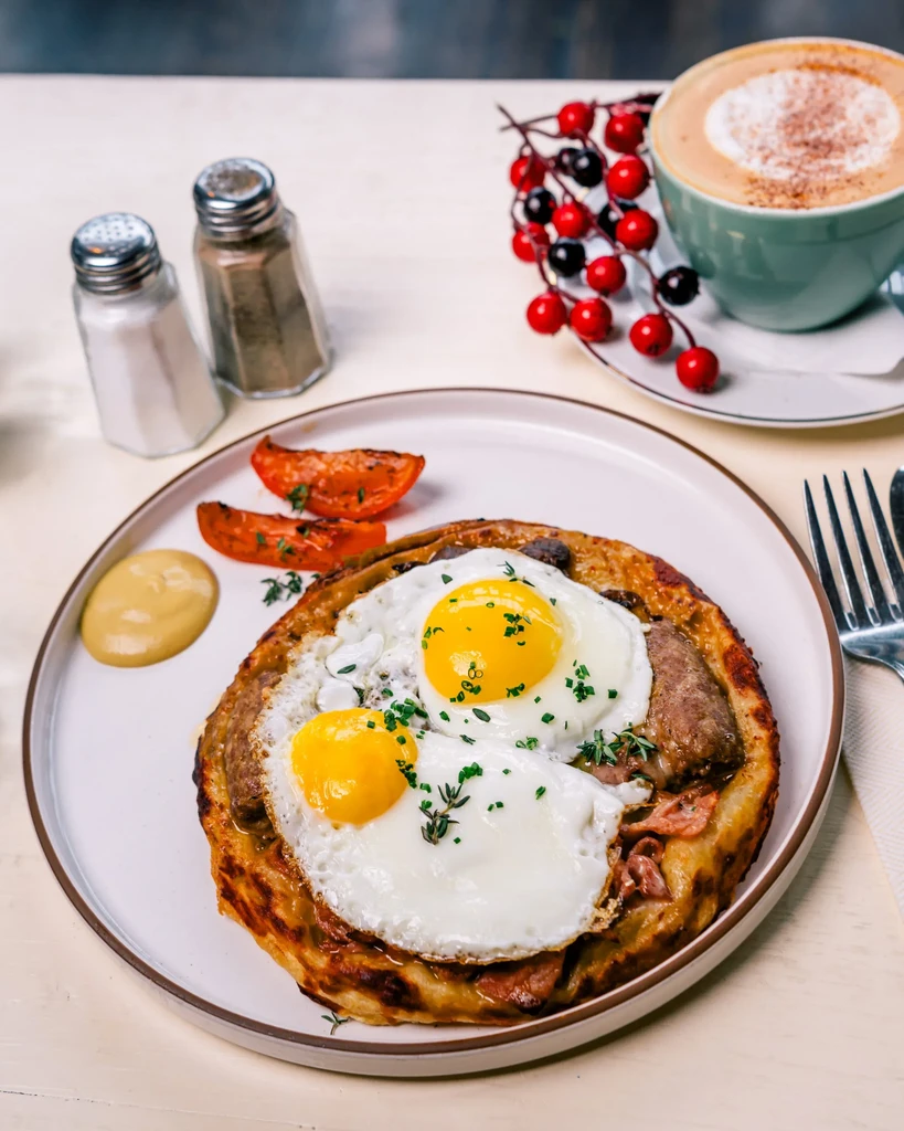 Breakfast plate with fried eggs, sausage, tomatoes, and a latte.