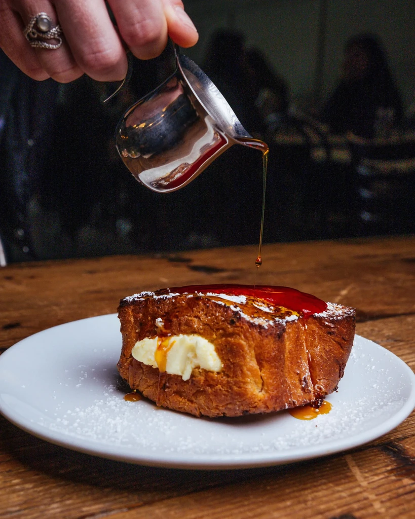 A hand pours maple syrup onto golden French toast with sugar.