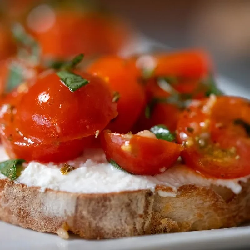 Toasted bread topped with creamy cheese, red cherry tomatoes, green herbs.