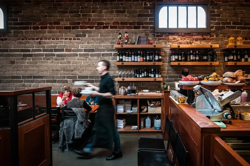 Busy rustic restaurant interior with exposed brick, customers, and food counter.