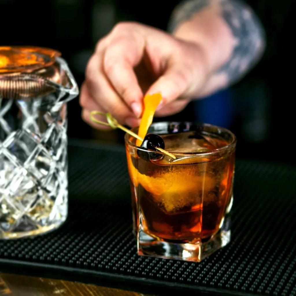 Bartender's tattooed hand placing orange peel in an Old Fashioned.