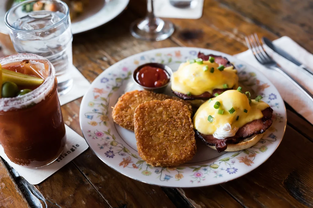 Close-up of Eggs Benedict, hash browns, Bloody Mary on rustic table.