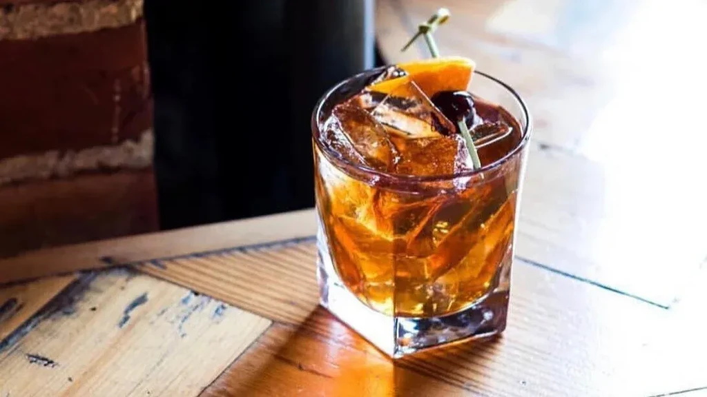 Amber-brown cocktail with orange slice, cherry, on a wooden table.