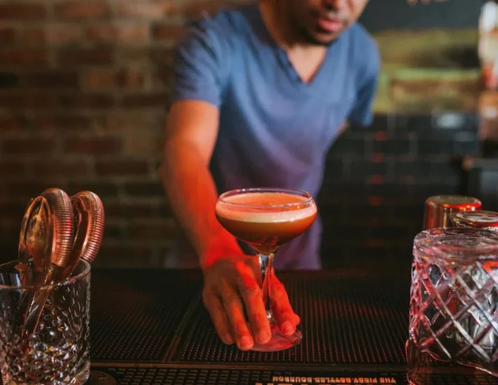 Bartender serves amber cocktail with foamy top in a coupe glass.