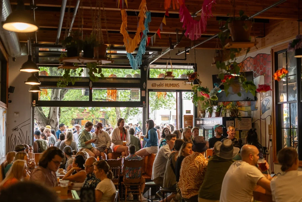 Crowded, vibrant restaurant with high ceilings, plants, and open doors.