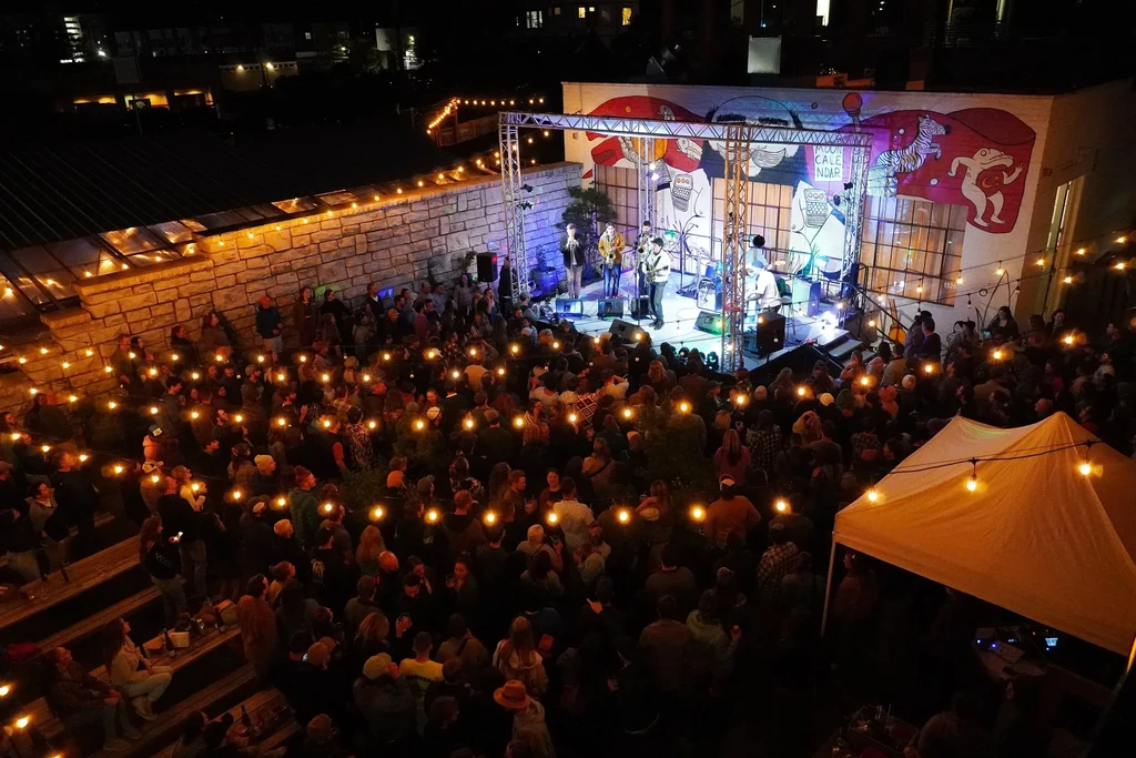 Outdoor evening concert with a band, large audience, and string lights.