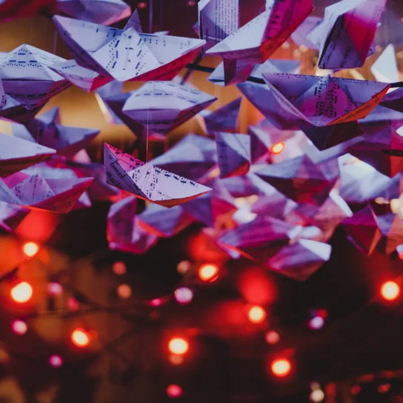 Suspended purple and pink origami paper boats, warm bokeh lights.
