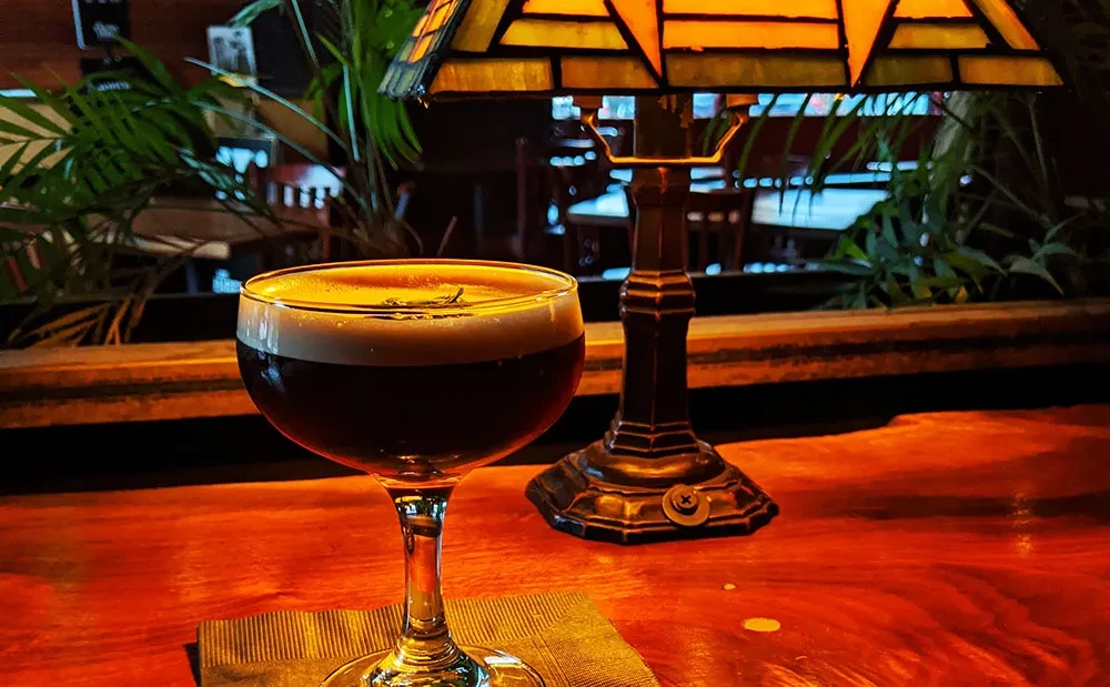 Amber cocktail in coupe glass with green sprig, beside Tiffany lamp.