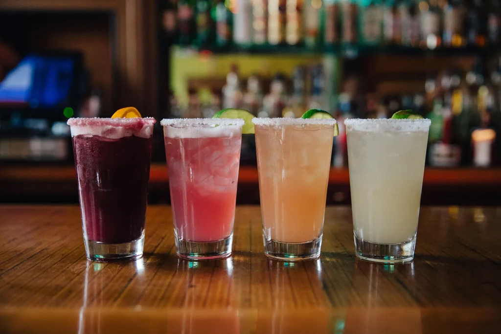Four colorful, garnished cocktails on a reflective wooden bar.