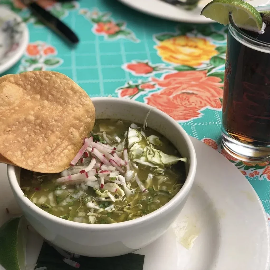 Green soup with cabbage, radishes, tostada, and drink on floral tablecloth.