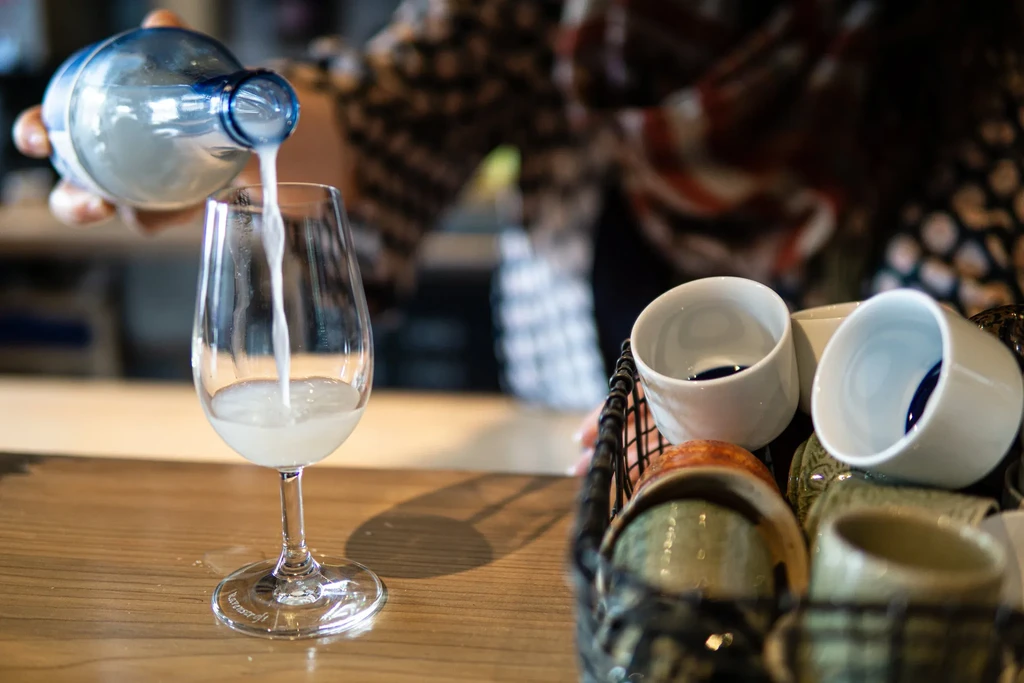 Person pours cloudy beverage from blue-capped bottle into wine glass.