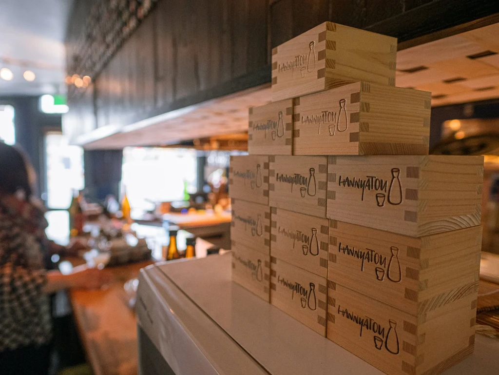 Cedar boxes with "HANN'YATOU" and sake logo stacked in a restaurant.