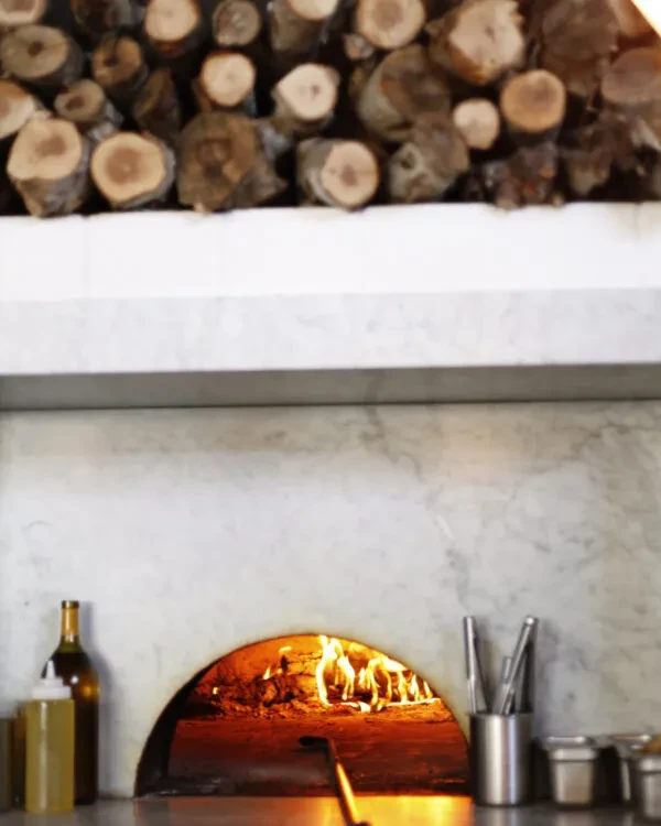 Wood-fired oven with active flames and stacked firewood in a kitchen.