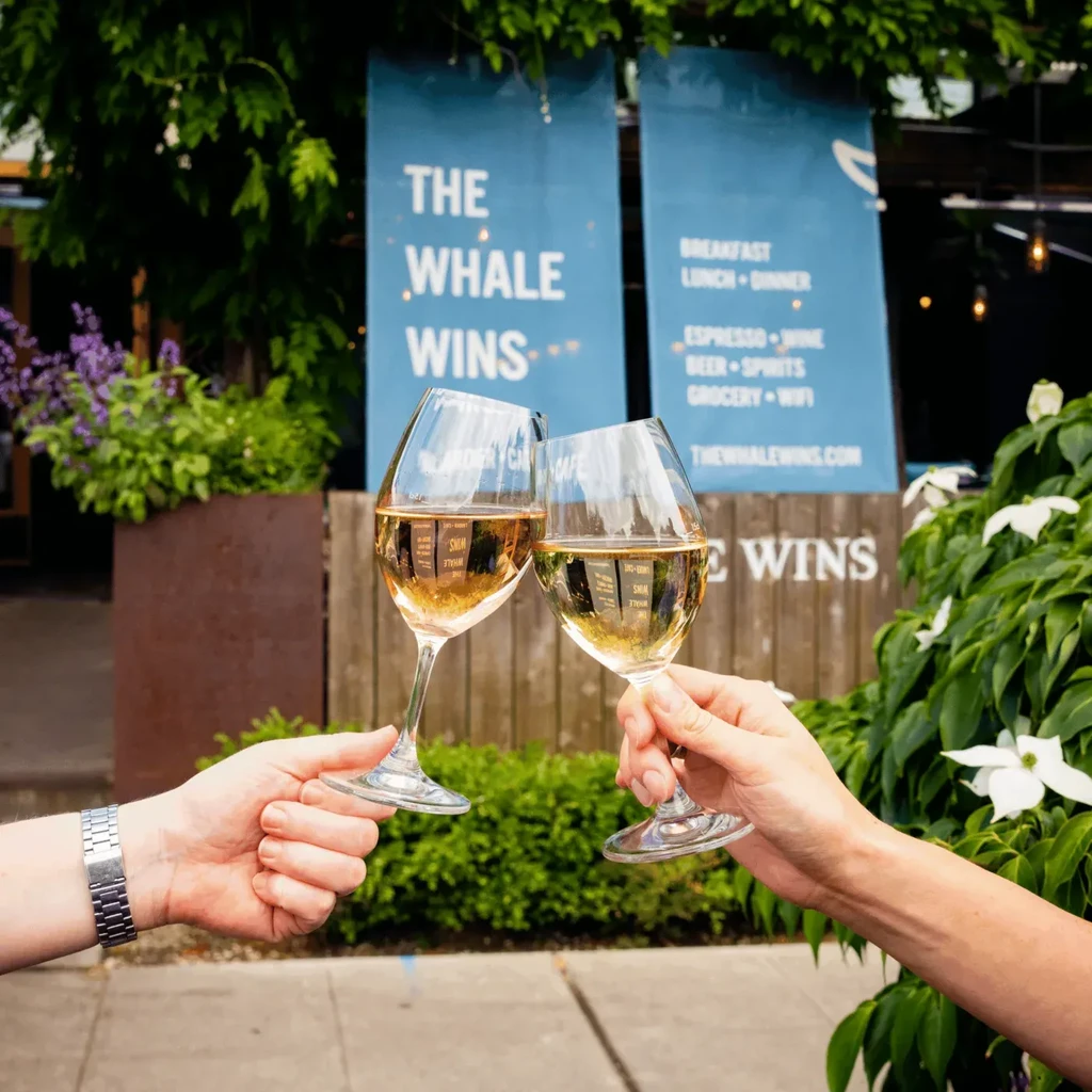 Hands toast wine glasses outdoors; "The Whale Wins" banner visible.
