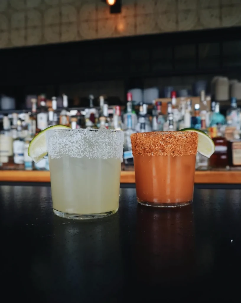 Two distinct cocktails with lime and unique rims on a bar counter.