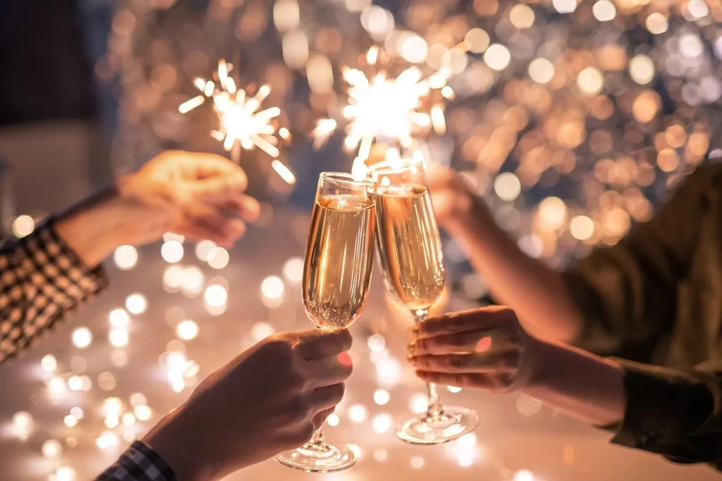 Two hands clink champagne flutes with sparklers in a festive glow.