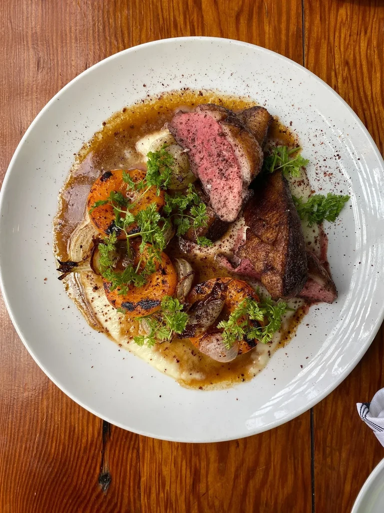 Overhead view of plated seared meat, grilled fruit, herbs, and sauce.