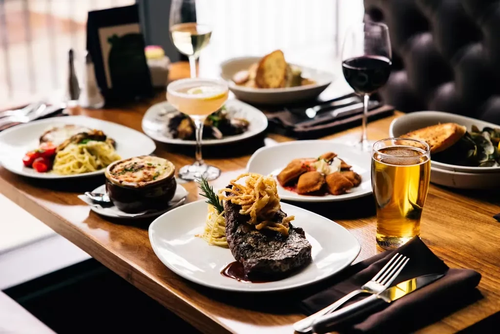 Plated steak, pasta, baked soup, and drinks on a restaurant table.