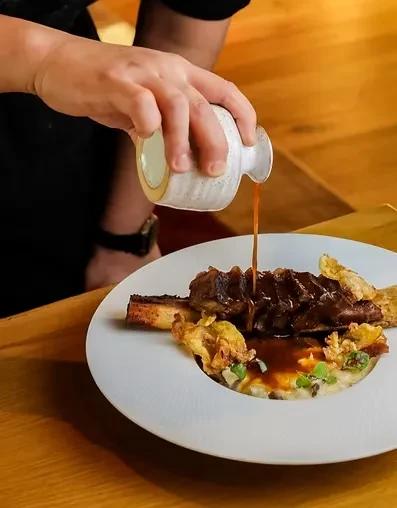 Hand pouring dark sauce over braised short rib with potatoes and greens.