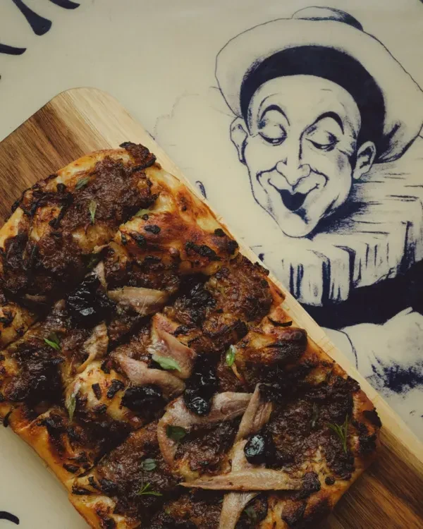 Sliced rectangular flatbread with anchovy, olive, herb toppings; jester art.