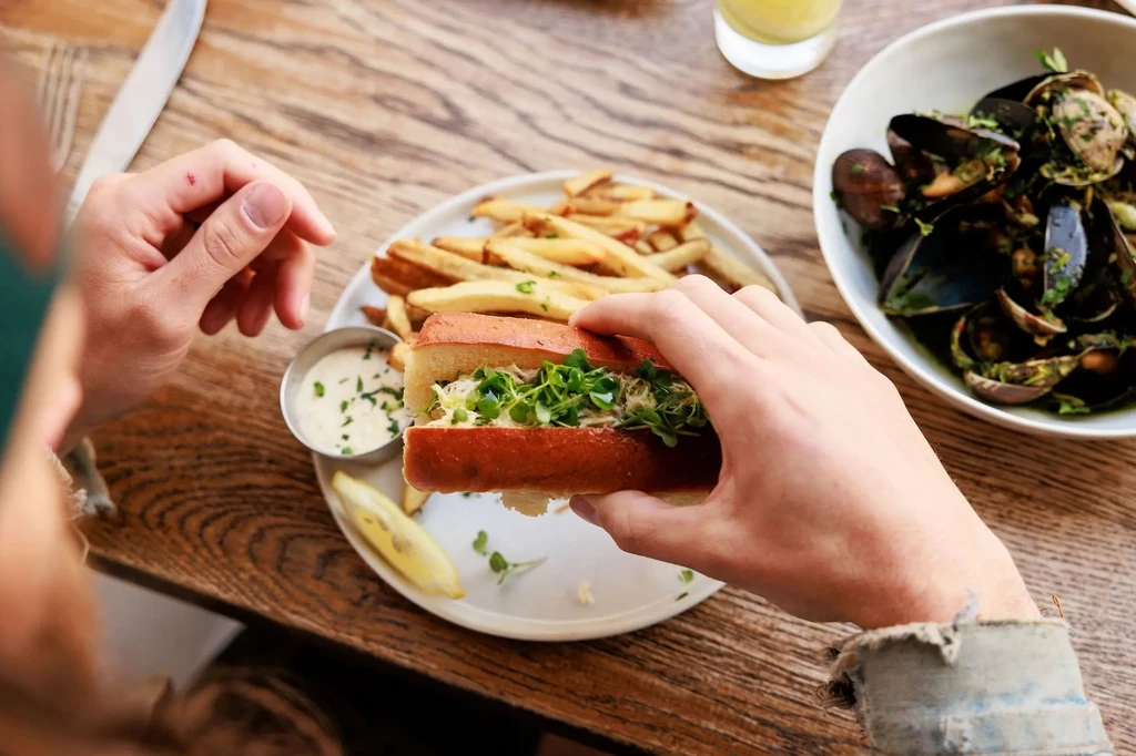 Hands holding seafood sandwich, fries, sauce, and mussels on a wooden table.