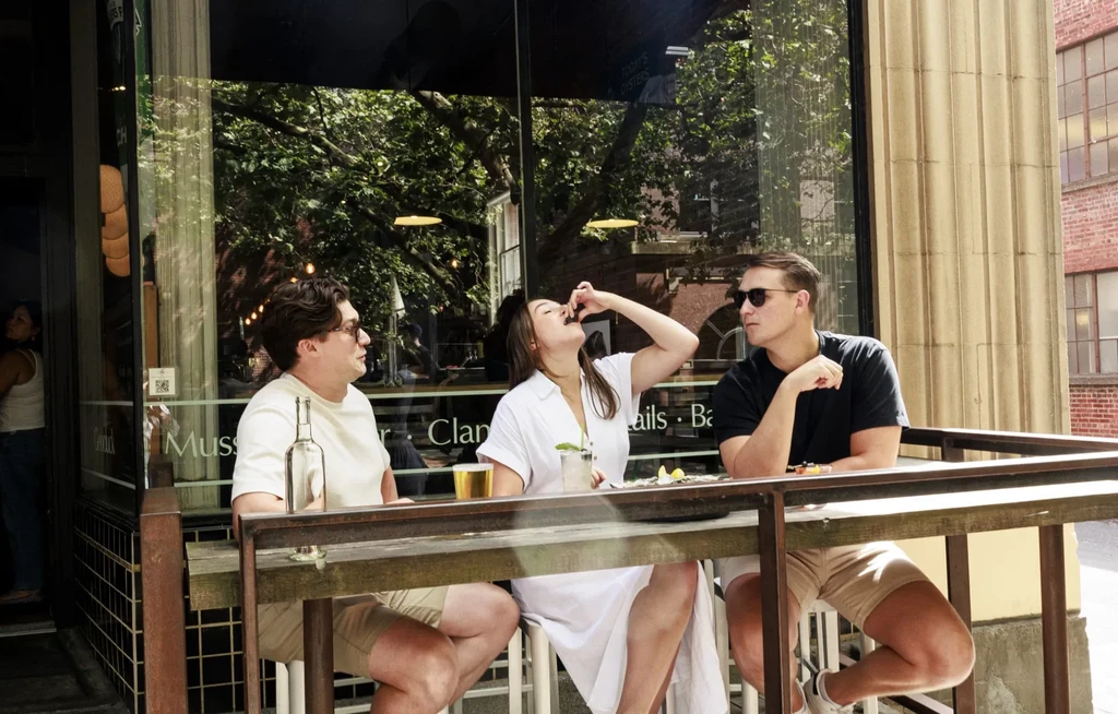 Three people eating oysters and drinks at a modern restaurant window.