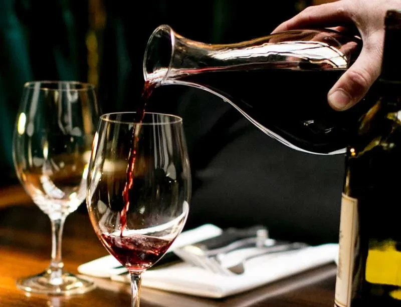 A hand pours red wine from a decanter into a wine glass.