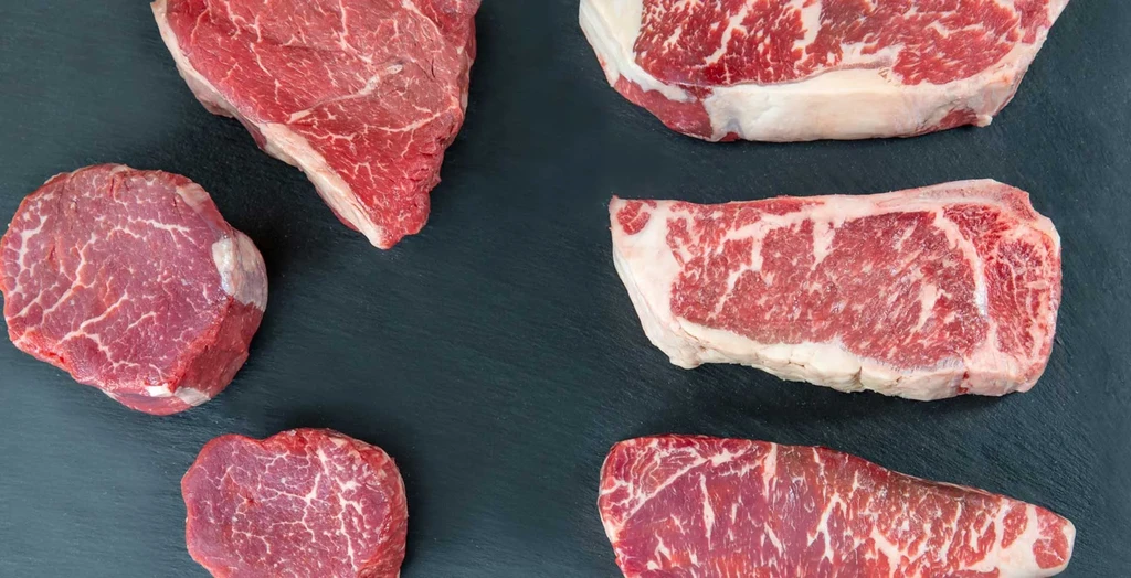Five raw beef steaks with marbling on a dark stone surface.