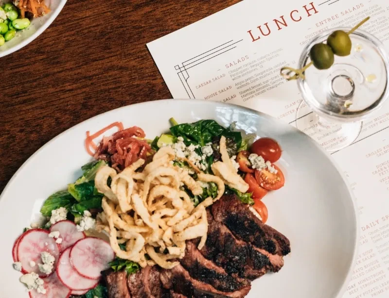 Grilled steak salad, martini, and menu on dark wooden table.