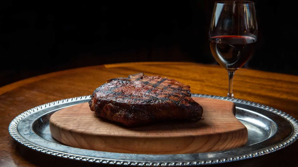 Grilled steak on wooden board with red wine glass in elegant setting.