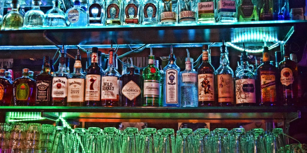 Blue and green lit bar shelves display liquor bottles and empty glasses.