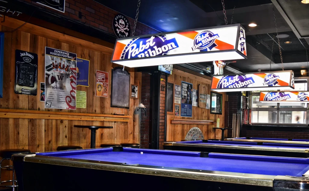 Rustic bar with three blue pool tables, wood walls, and PBR lights.