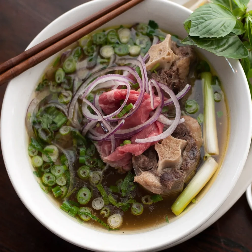 Bowl of Vietnamese pho with rare beef, oxtail, onions, basil, chopsticks.