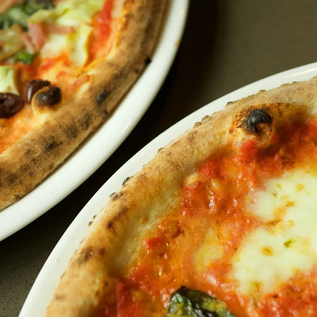 Foreground pizza with basil, background pizza with olives, on plates.