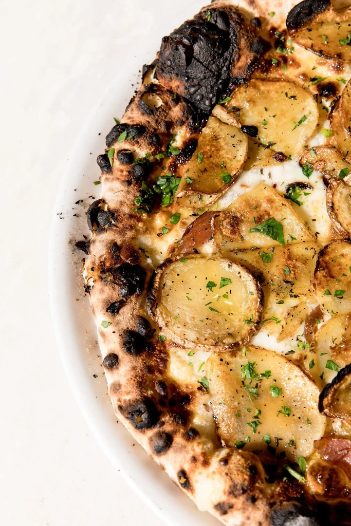 Close-up of gourmet potato pizza with charred crust and green herbs.