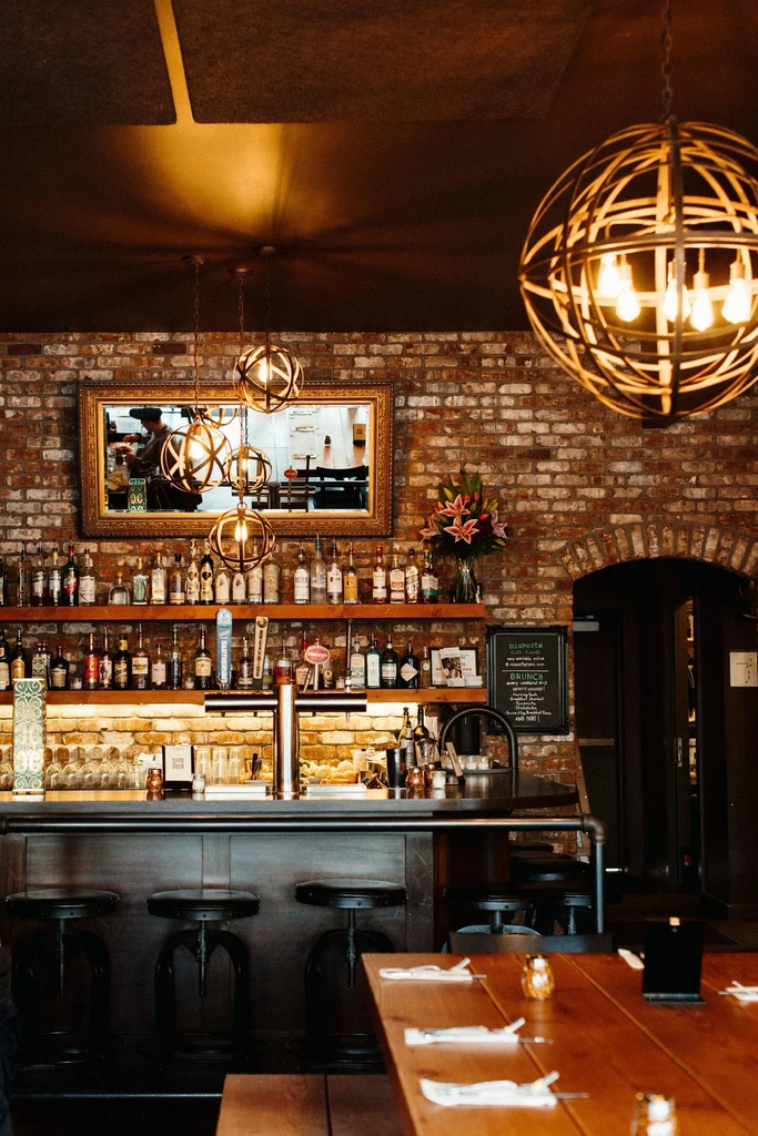 Warm, rustic-industrial bar interior with exposed brick and illuminated liquor bottles.